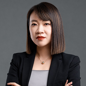 Portrait of Dan Zhang, a woman with straight, chin-length dark hair and red lipstick, wearing a black blazer over a gray top and a thin pendant necklace, arms crossed, against a gray background.