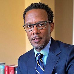 Portrait of a man with short dark hair and a graying goatee, wearing rectangular glasses, a navy blazer, light blue shirt, and a navy striped tie.