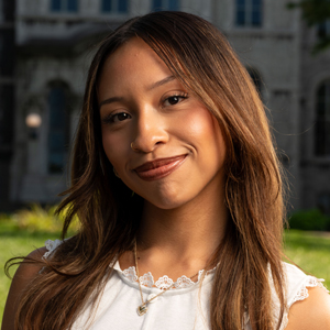 A student smiles with long brown hair, wearing a white top with lace trim.