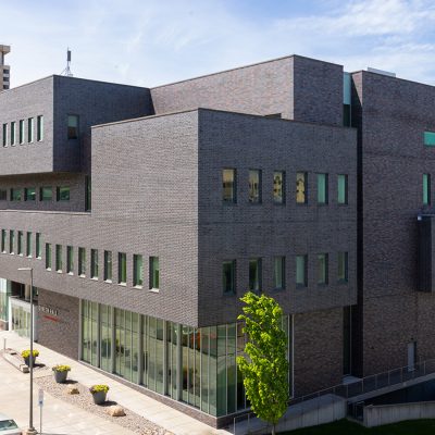 Dineen Hall, a modern dark brick building with stacked, offset rectangular volumes, rows of narrow green-tinted windows, and a glass-walled ground floor, seen from a corner on a sunny day.