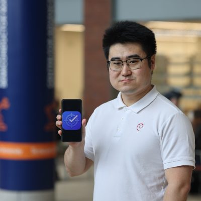 A person holds up a smartphone showing a blue app icon with a checkmark and clock, standing in a busy campus building with a blue column