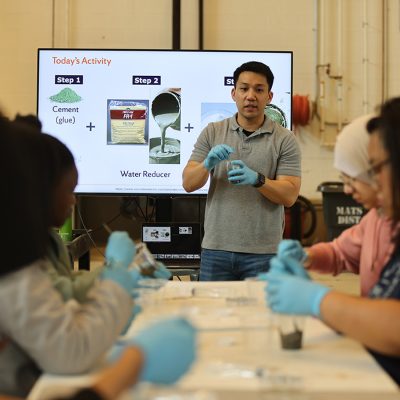 A professor in a gray polo and blue gloves holds a beaker and speaks to participants at a lab table, in front of a screen reading “Today’s Activity” with steps for mixing cement and a water reducer.