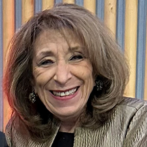 Portrait of Evelyn Granieri, a smiling woman with shoulder-length brown hair and drop earrings, wearing a metallic gold jacket over a dark top, in front of a wood-paneled wall.