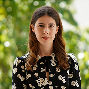 Headshot of Fitore Hyseni, a woman with long auburn hair and small gold hoop earrings, wearing a black blouse with a cream floral pattern, standing in front of blurred green foliage.