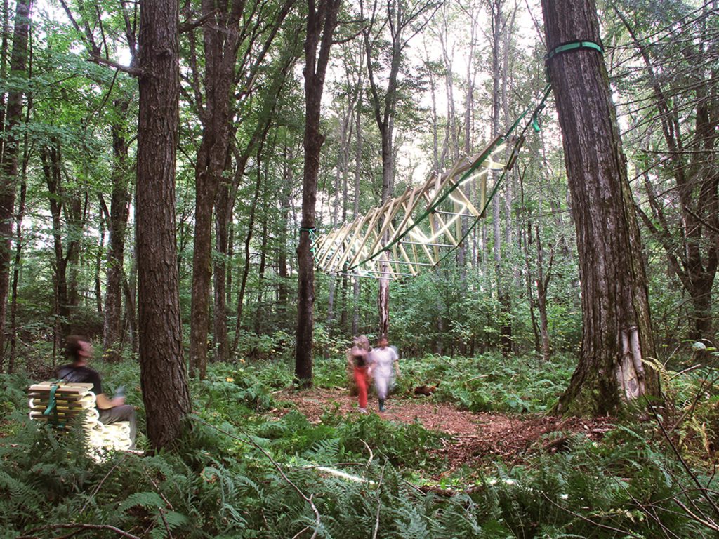 A lightweight lattice of pale wooden slats hangs suspended between trees in a green forest, with two blurred figures walking beneath it and another person seated on a wooden bench at left.