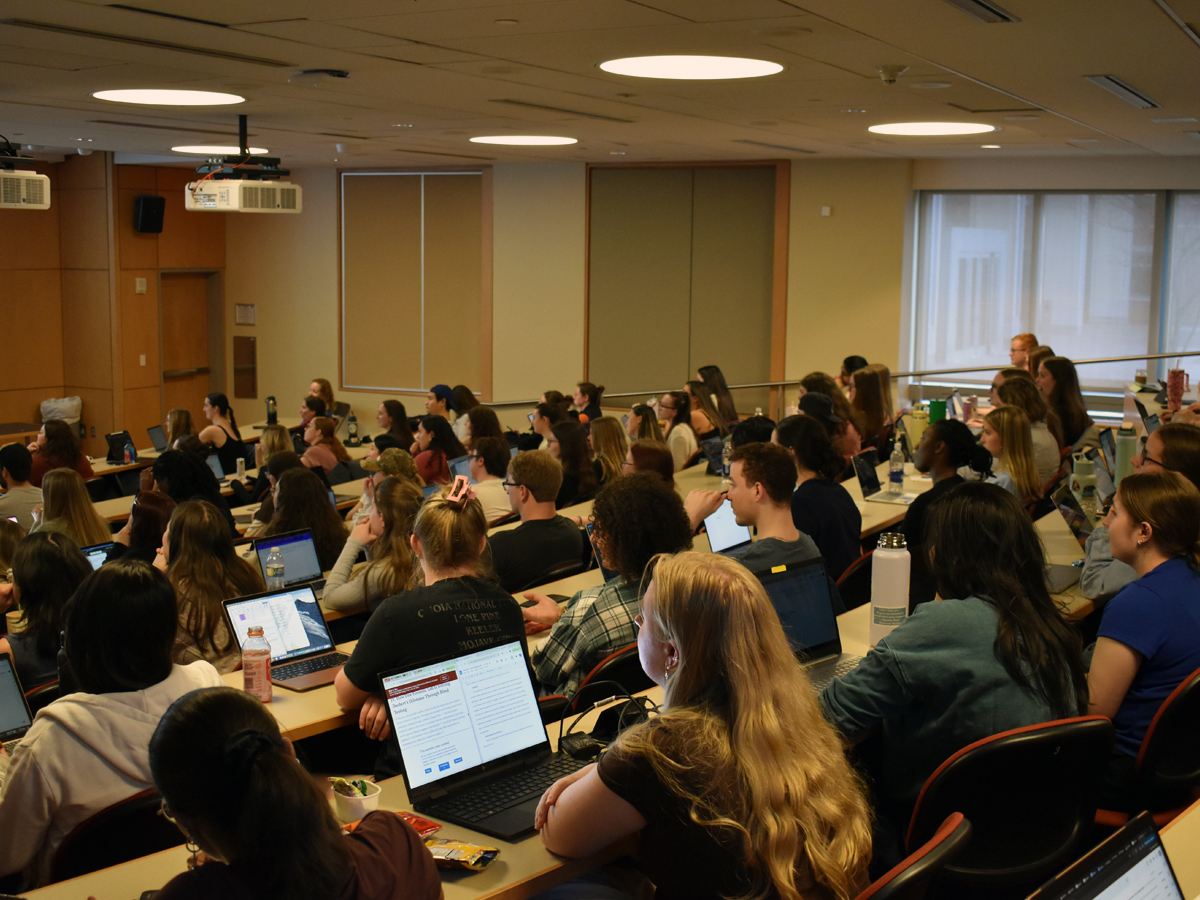 Students seated in a tiered lecture hall, many with open laptops, facing the front of the room.