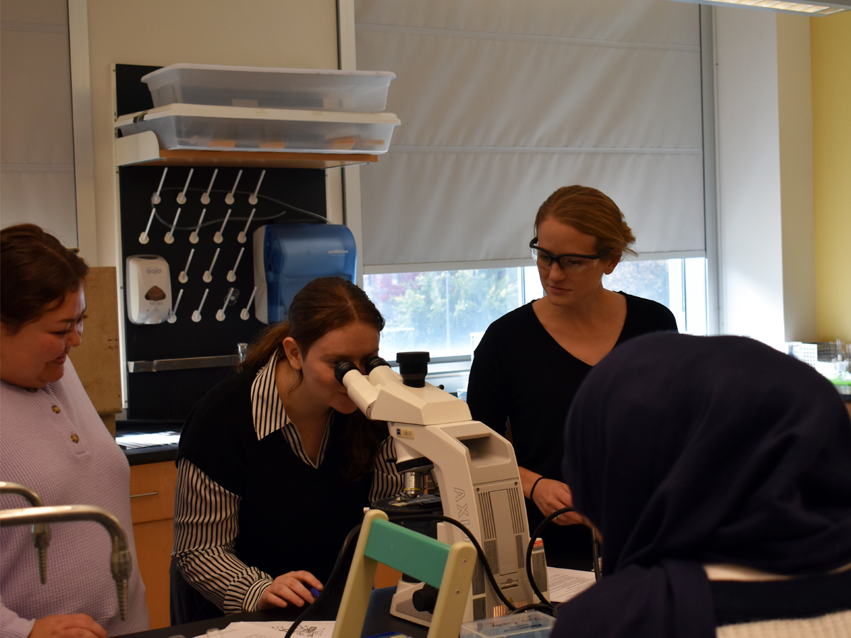 A student looks through a microscope as classmates and an instructor watch in a lab.