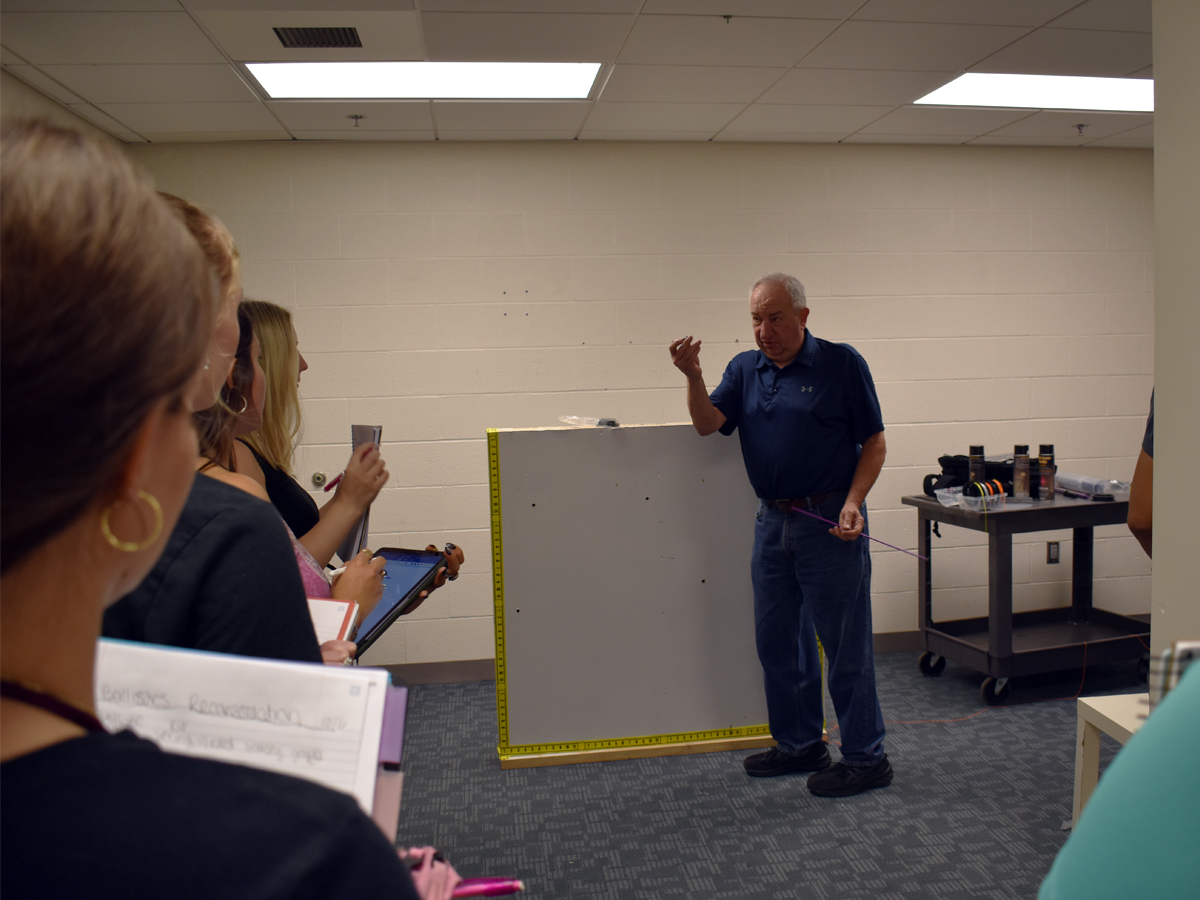 An instructor gestures beside a measured panel while students with notebooks and tablets watch.