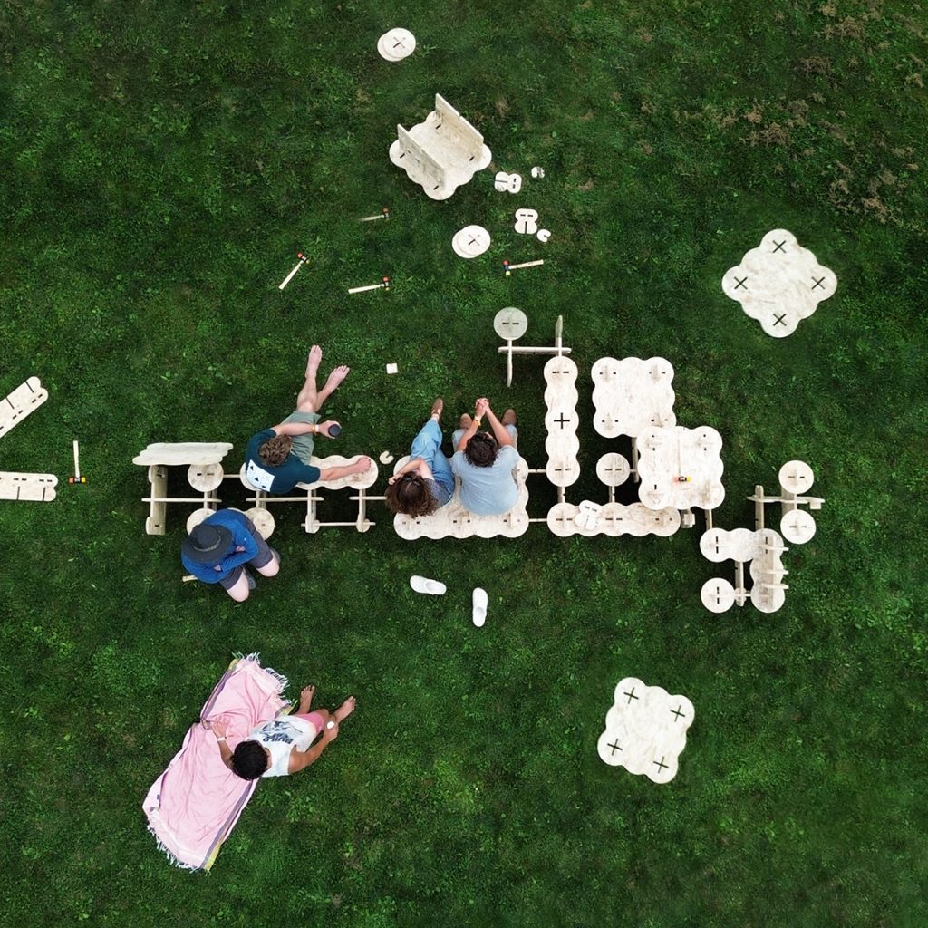 Aerial view of five people on a lawn around a modular plywood bench with scalloped, cloud-like edges, with loose cut pieces and mallets scattered across the grass.