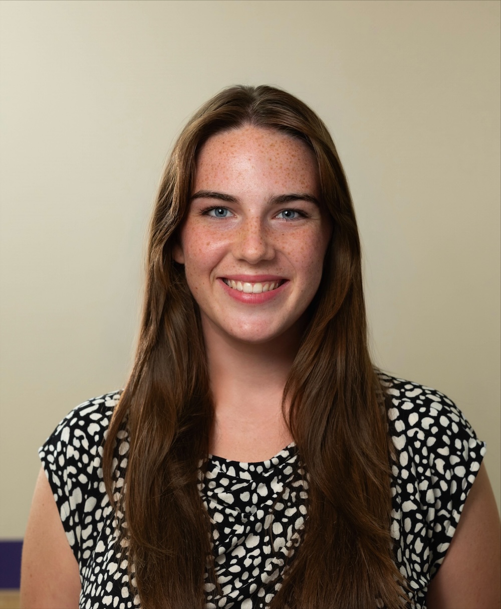 Professional headshot of a person with long brown hair wearing a black-and-white patterned top against a neutral background.