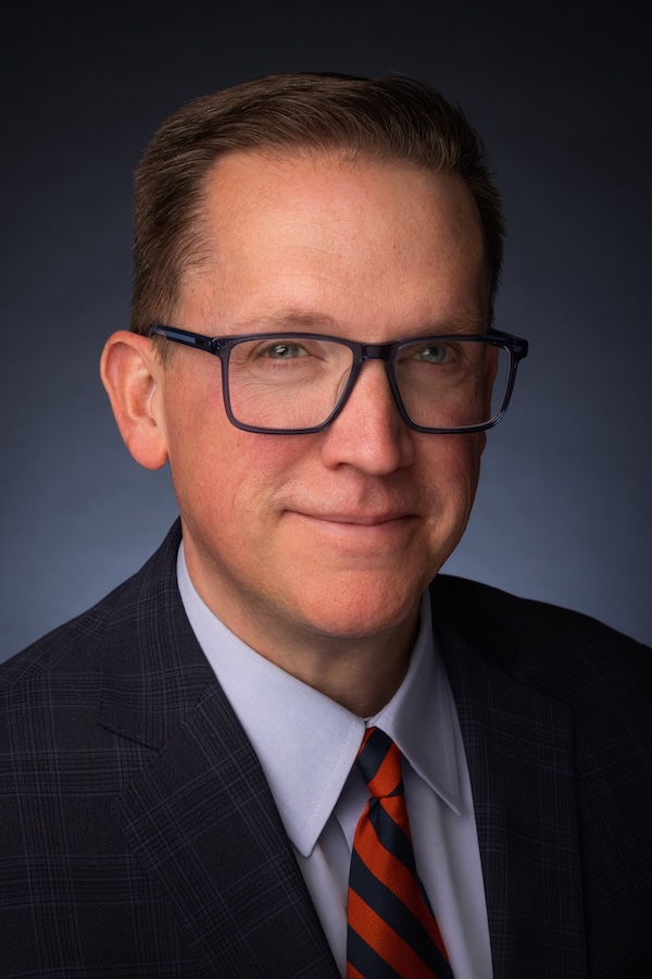 Professional headshot of a person wearing glasses, a dark plaid suit jacket, light dress shirt, and striped orange-and-navy tie against a dark blue background.