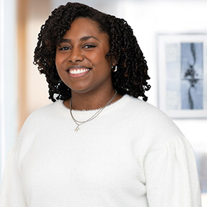 Portrait of Hailey Williams, a smiling woman with shoulder-length twisted curly hair, wearing a white knit sweater and a delicate silver necklace with a small pendant, in a bright room with a framed picture behind her