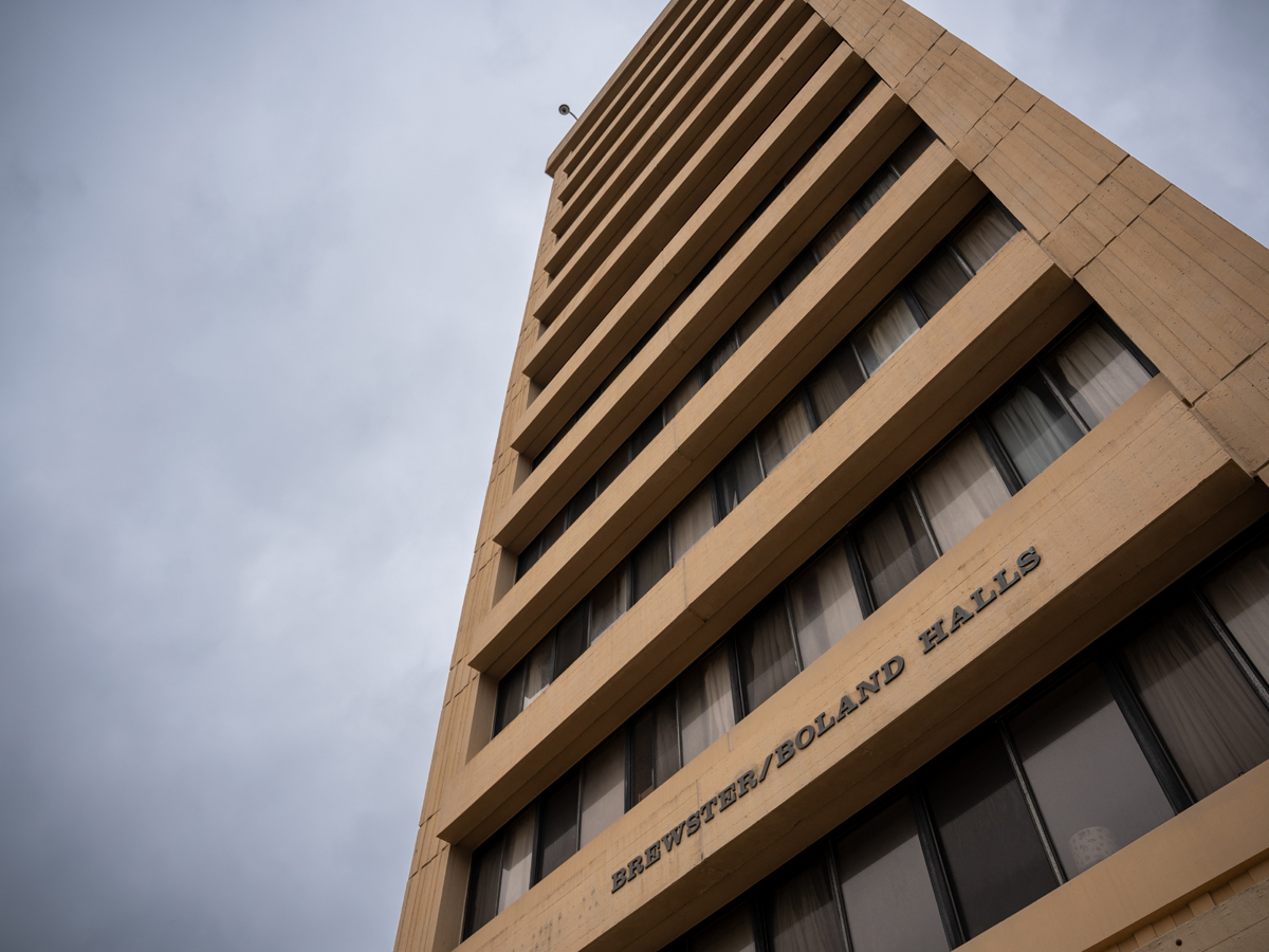 Looking up at the tan concrete facade of Brewster/Boland/Brockway Halls under a cloudy sky.