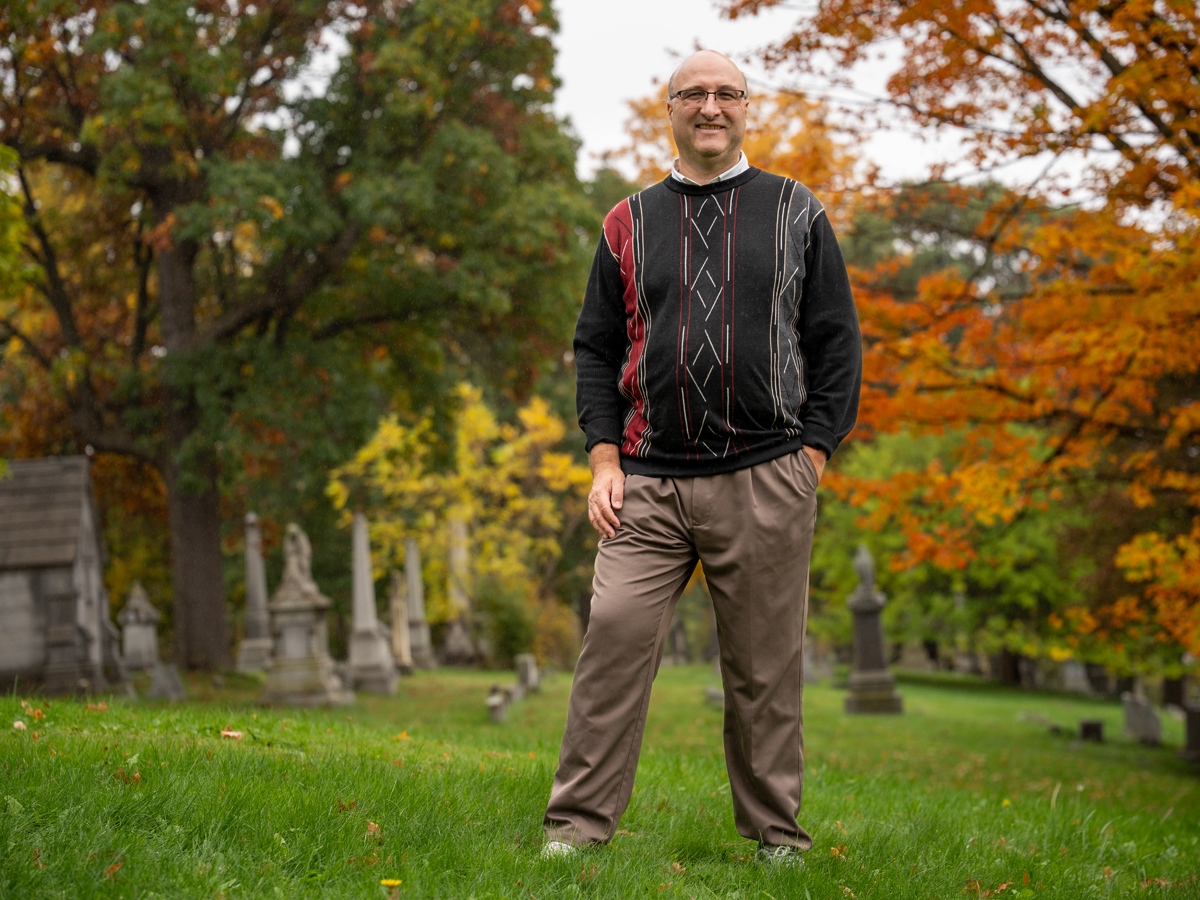 Man in glasses and a patterned sweater stands smiling among gravestones, surrounded by fall foliage.