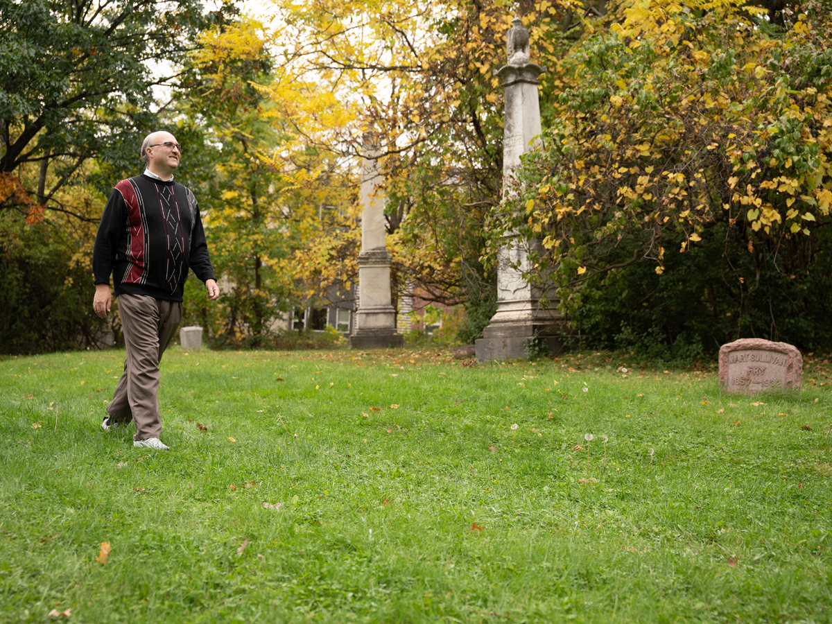 Man in a patterned sweater walks through a grassy cemetery with obelisks and autumn trees.