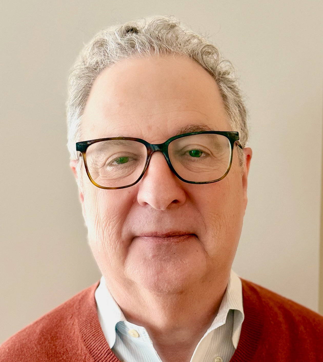 Close-up headshot of a person with short gray hair, glasses, and a rust-colored sweater.