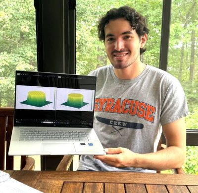 A smiling young man in a Syracuse Crew T-shirt holds up a laptop showing two 3D graphs of a block-shaped surface, seated at a wooden porch table with trees behind him.