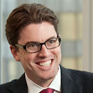 Portrait of Ian Schuman, a smiling man with swept-back brown hair and dark rectangular glasses, wearing a dark suit, white shirt, and red patterned tie, in front of a softly blurred window background