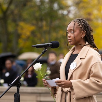 Woman in a camel coat speaks at a microphone, holding a white rose, at an outdoor autumn memorial gathering.