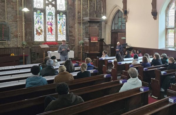A speaker addresses a group seated in the pews of a historic stone church with stained glass windows.