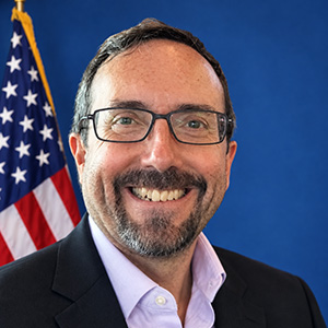 Portrait of John R. Bass, a smiling man with glasses and a salt-and-pepper beard, wearing a dark blazer and open-collar light shirt, in front of a blue background and an American flag