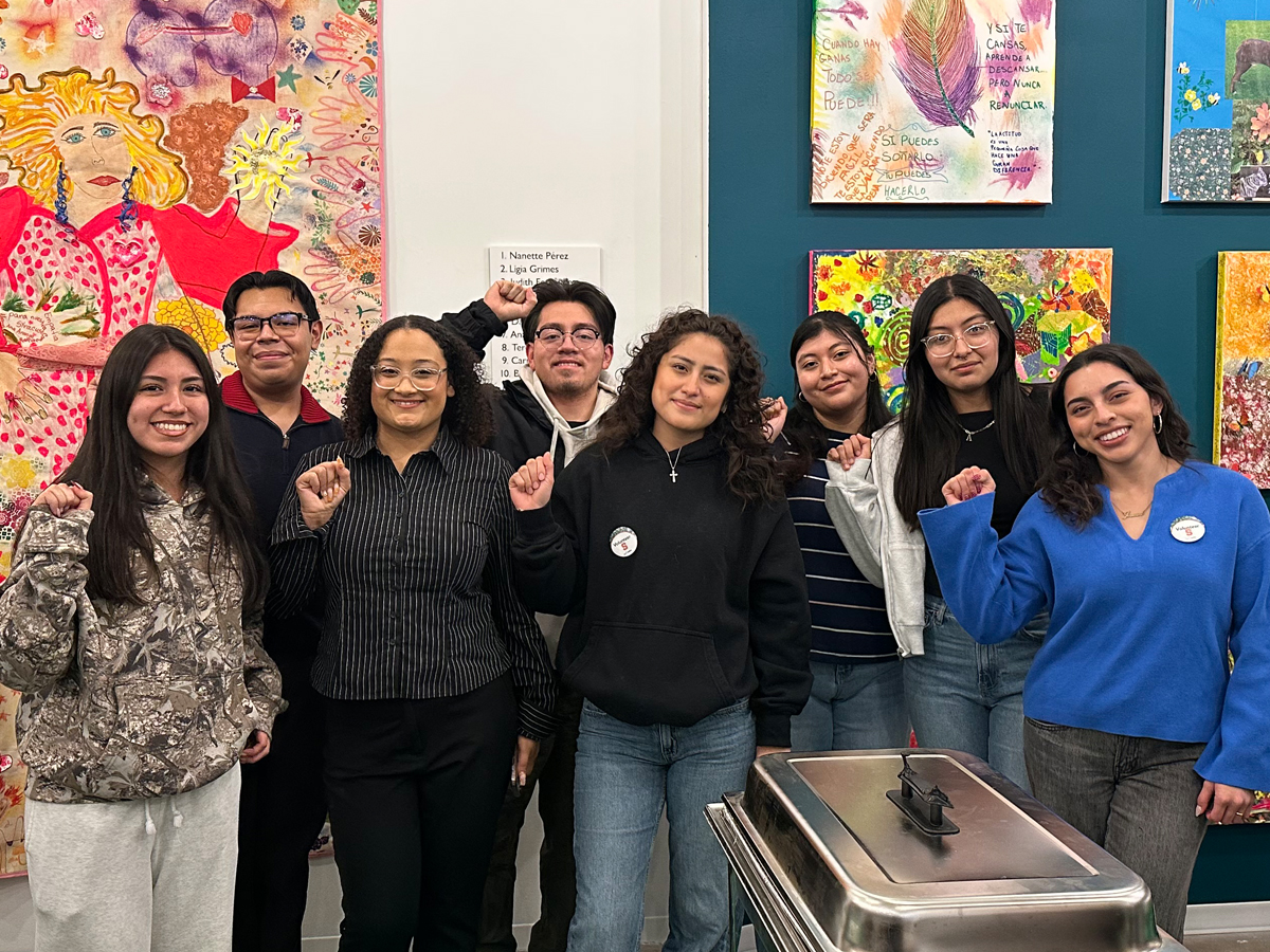 Eight students raise fists in front of colorful artwork, with a food warmer in the foreground.