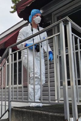 A person in a protective suit, mask and gloves holds a camera on a porch with metal railings.