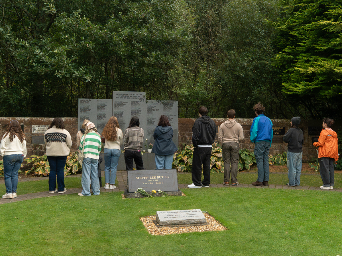 Visitors stand before a stone memorial wall in a garden, with flowers laid at a marker.
