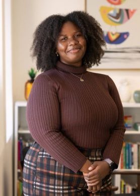 A smiling woman in a brown turtleneck and plaid pants stands in front of bookshelves and colorful abstract artwork.