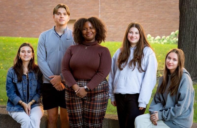 Five smiling people pose together on a lawn in front of a brick building, some standing and some seated on a low wall.