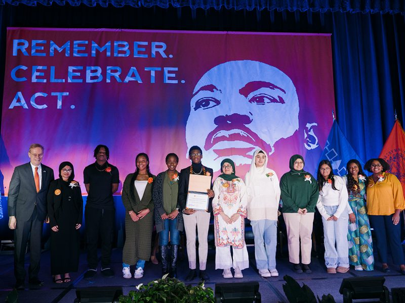 Twelve people pose on stage with a certificate beneath a banner reading “Remember. Celebrate. Act.” with an MLK portrait.