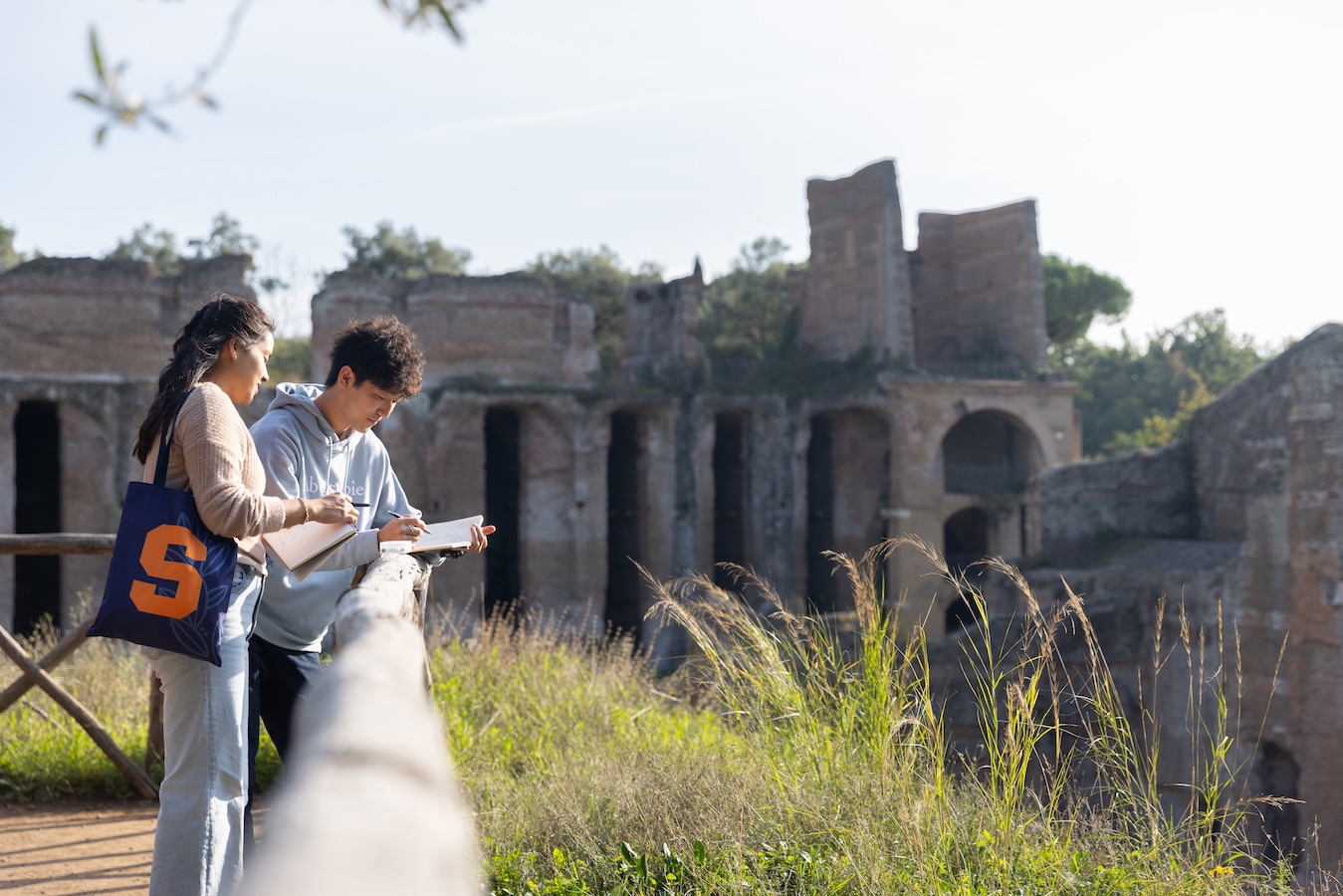 Two students sketch and take notes while observing ancient ruins from a walkway overlook.