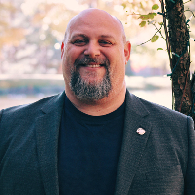 A person smiles outdoors in a gray blazer and navy shirt with a small lapel pin.