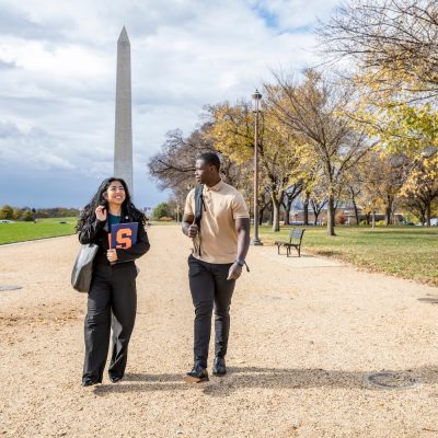 Two students walk along a path near the Washington Monument, with autumn trees and a cloudy sky in the background.