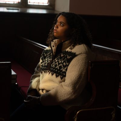 Woman in a fleece sits in a church pew, gazing upward as light falls across her face.