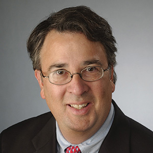 Headshot of Peter Blanck, a smiling man with dark, graying hair and thin round glasses, wearing a dark suit jacket, light blue collared shirt, and red patterned tie against a gray background.