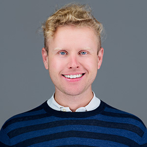 Portrait of Phil Porter, a smiling man with short, curly blond hair and blue eyes, wearing a navy and blue striped sweater over a white collared shirt, against a gray background