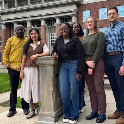Six people smile while posing for a photo outside of the Maxwell School.