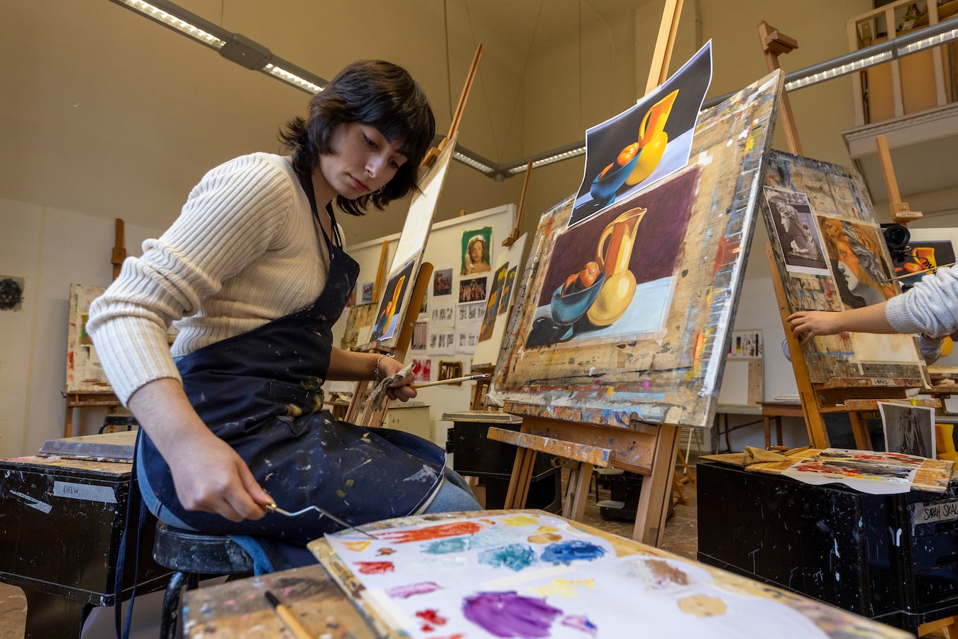 Person in a painting studio mixes colors on a palette while working at an easel displaying a still-life painting.
