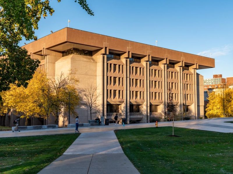 Modern concrete campus building with a tree-lined walkway and autumn foliage in warm evening light.
