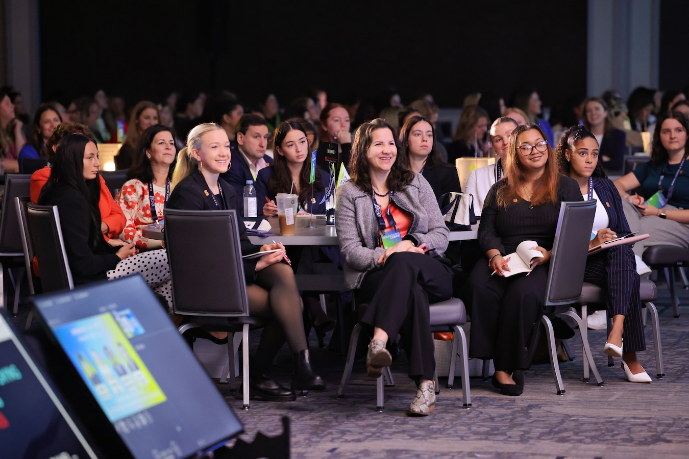 Attendees seated at round tables listening to a speaker during a conference session.