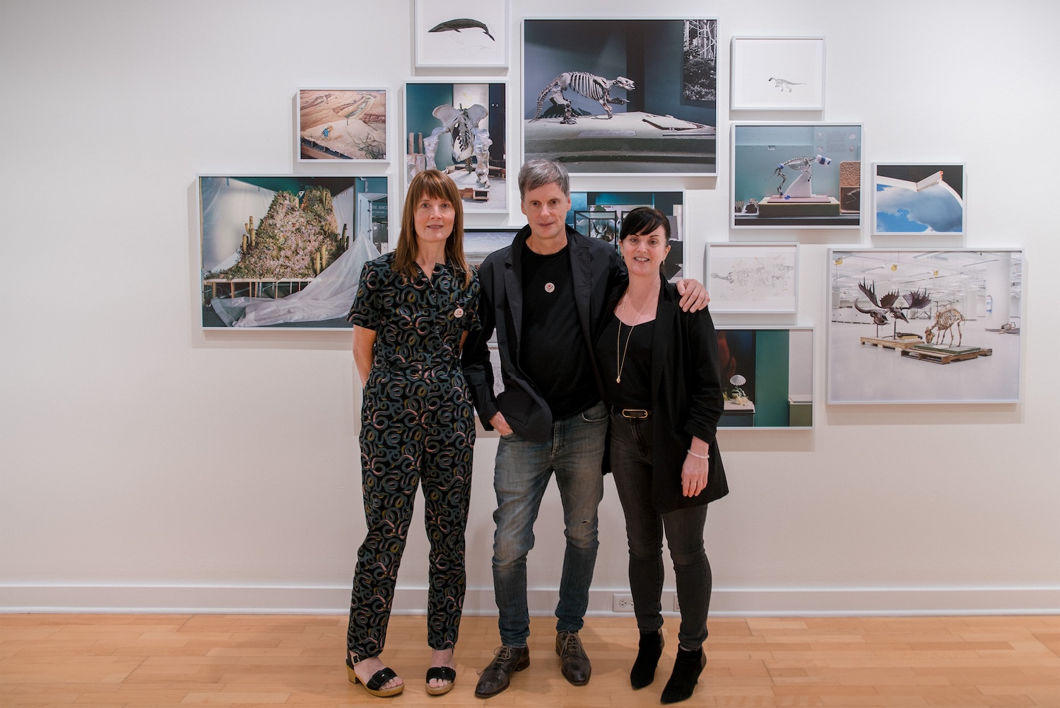 Three people pose in front of a gallery wall displaying framed photographs of museum and natural history exhibits.