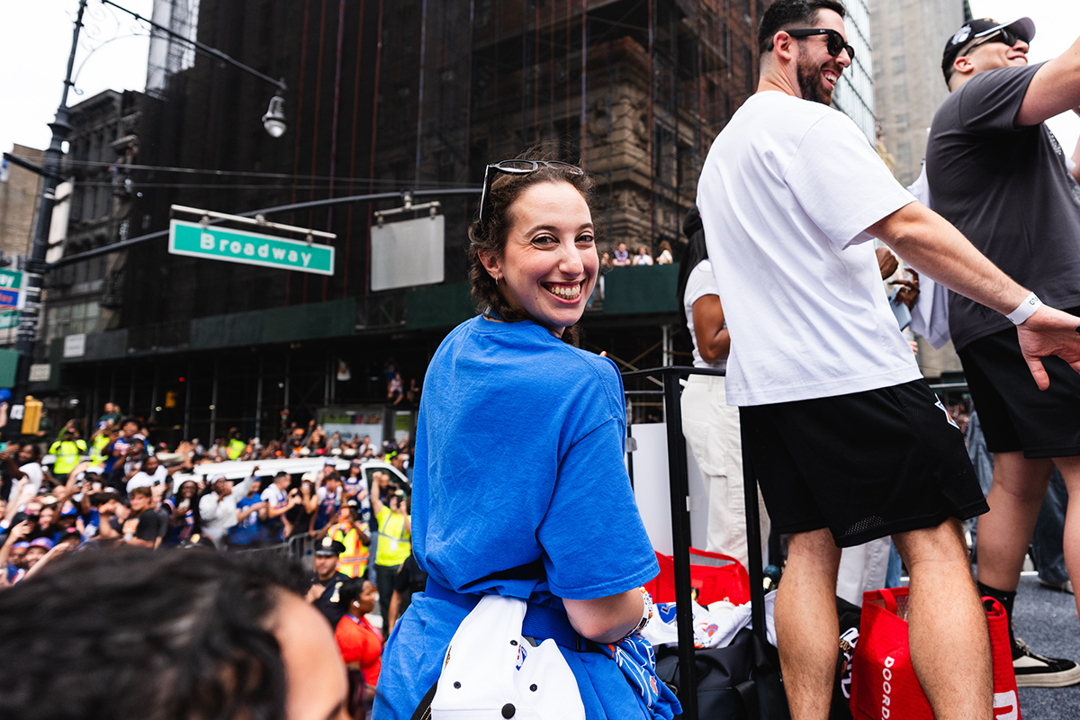A smiling person in a blue shirt rides a parade float on Broadway as fans watch.