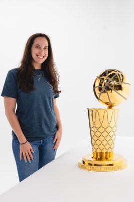 A smiling woman stands beside the gold NBA championship trophy against a white backdrop.