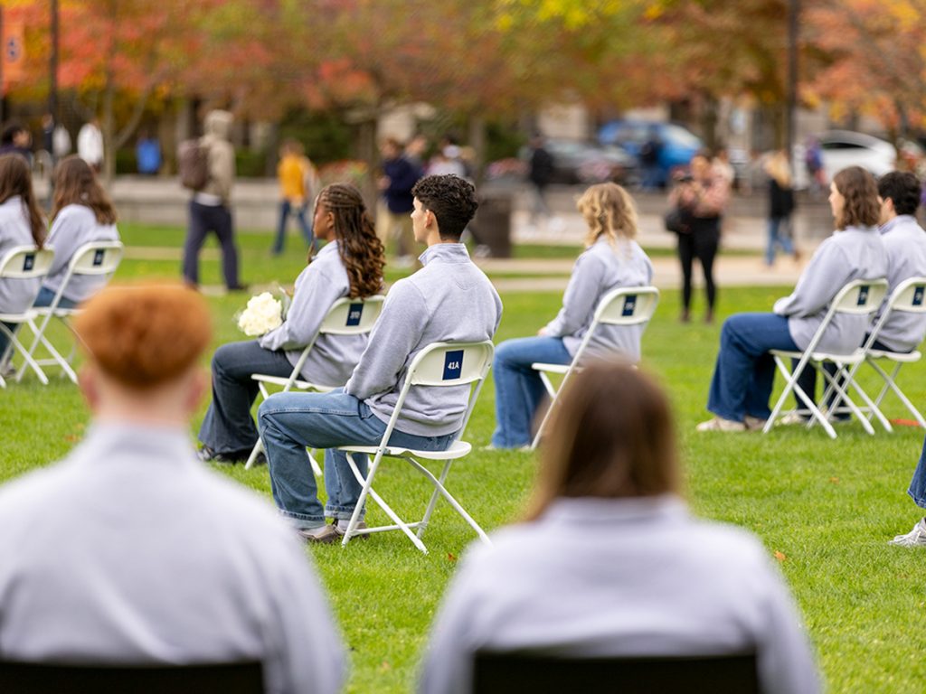 Students in matching light gray quarter-zip pullovers sit in white folding chairs on a grassy lawn, facing away from the camera, with autumn trees and passersby blurred in the background.