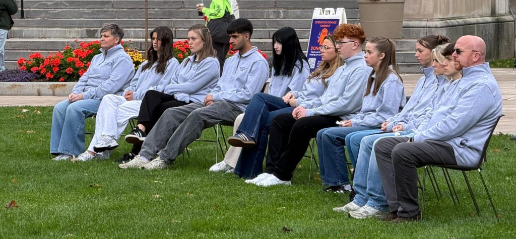 A group of about a dozen people in matching gray quarter-zip pullovers sit in rows of folding chairs on a lawn, looking off to the left with serious expressions. Red flowers and stone steps are behind them.