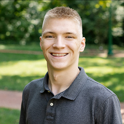 Headshot of a person in a gray polo shirt standing outdoors with greenery in the background.