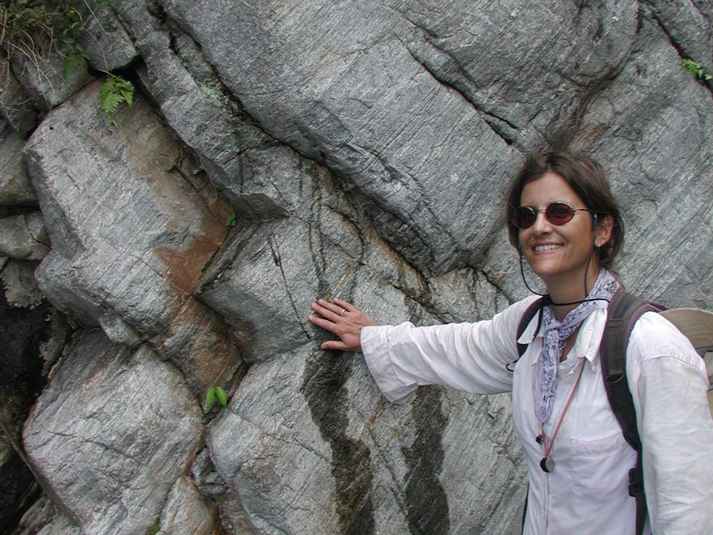 Smiling woman in sunglasses, a white shirt, and a patterned bandana rests her hand on a gray, layered rock outcrop with fine streaking and rust-colored staining.