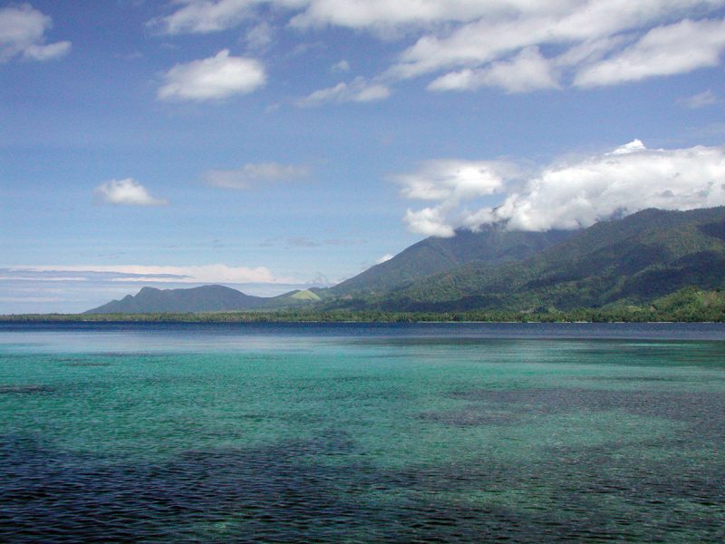 Turquoise tropical bay with dark blue patches over reefs, backed by forested green mountains with clouds resting on the peaks under a bright blue sky.