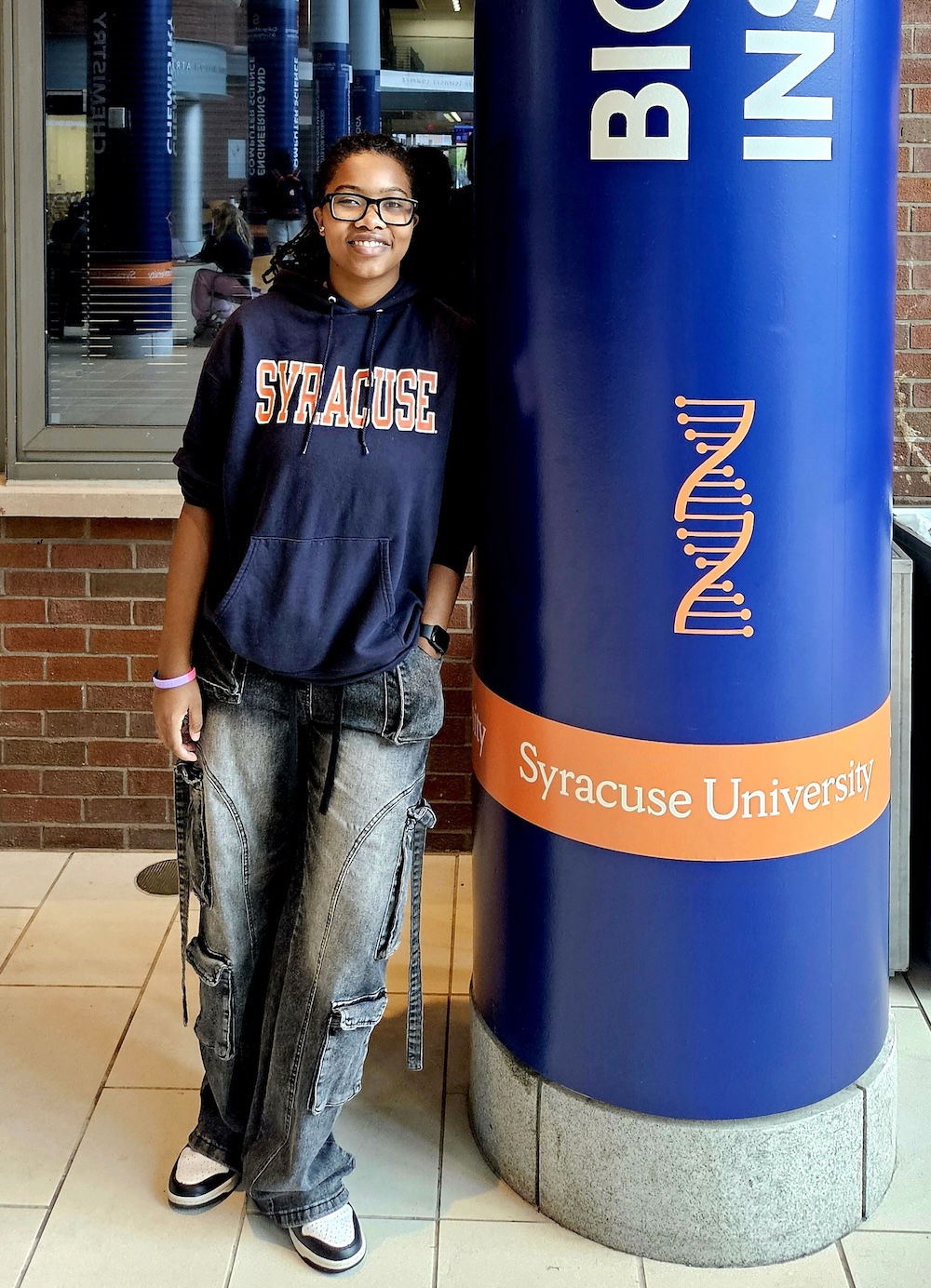 Person in a Syracuse University sweatshirt standing beside a campus pillar labeled “BIO” with a DNA graphic.
